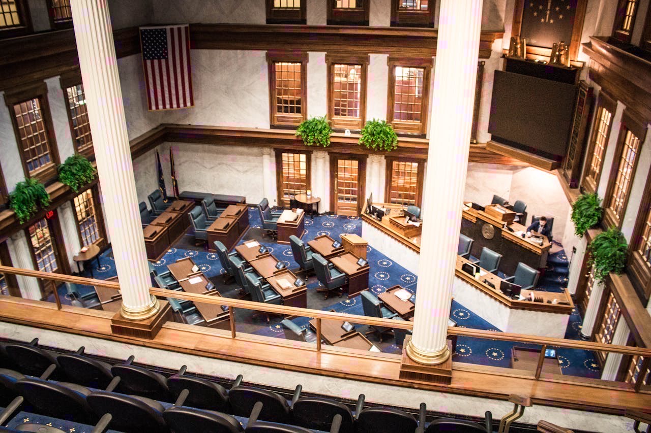 A state capitol building interior with legislative chamber desks and AI-themed documents, overhead shot, documentary style