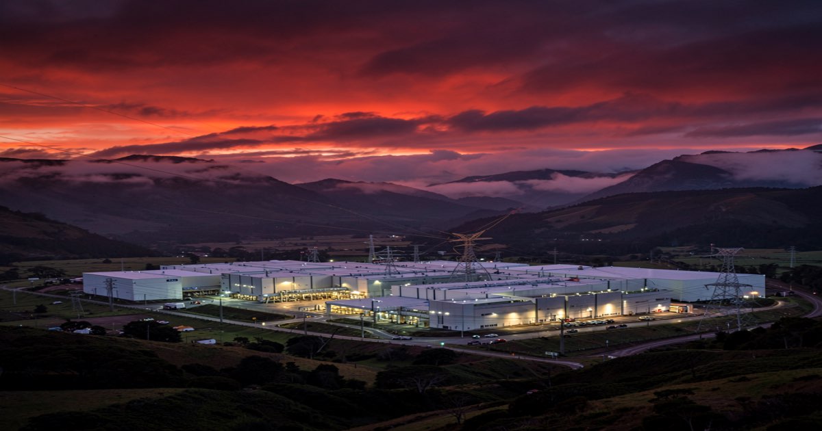 A massive data centre complex at dusk in a Southland landscape, power transmission lines converging on the facility, hydroelectric mountains in the background, ominous red and orange sky, no text no logos no letters no signs
