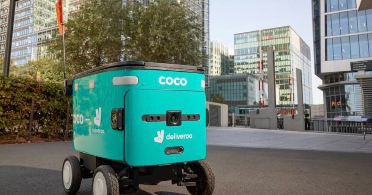 A turquoise Coco sidewalk delivery robot with Deliveroo branding and an orange warning flag on the pavement at Canary Wharf, London