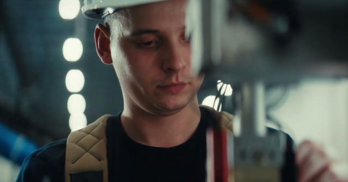 A worker in a hard hat and fall harness watches a robotic arm work inside Promise Robotics' production facility, a frame from the company's factory video.