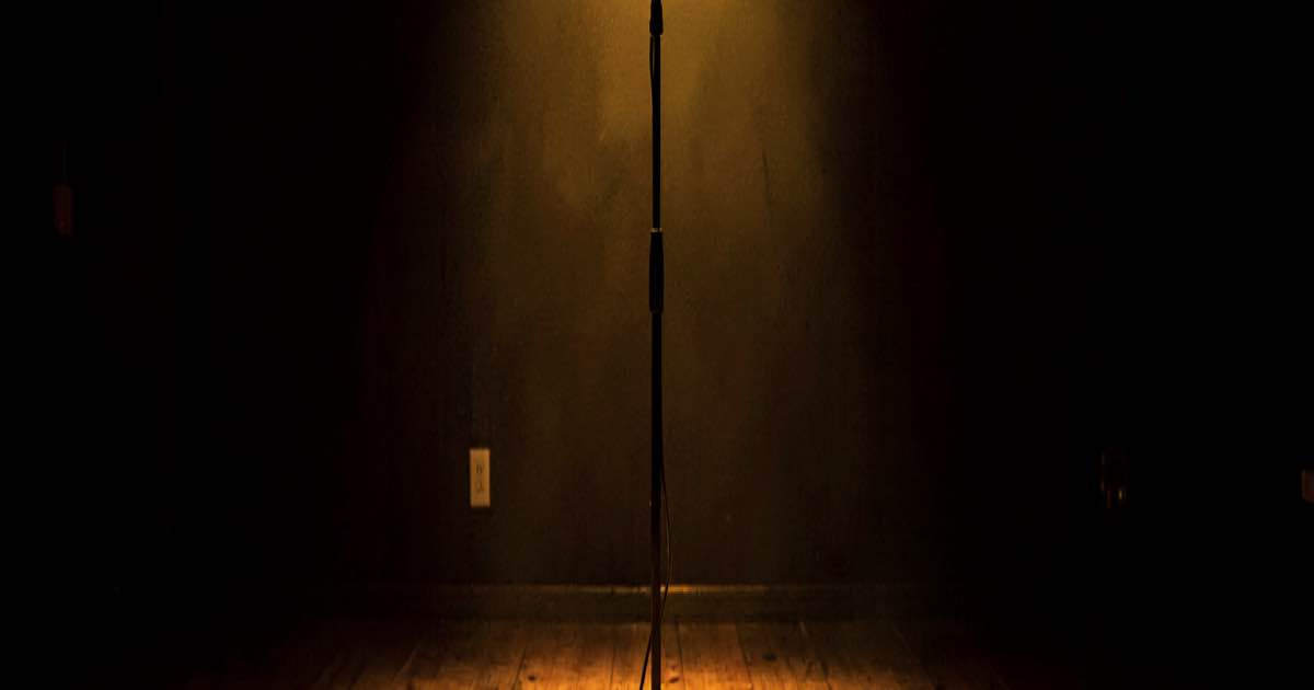Empty concert stage with a single microphone stand under warm spotlight, sheet music scattered on the floor, documentary style