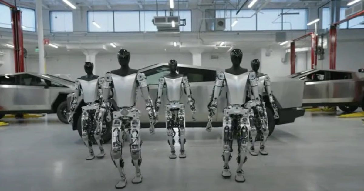 Five Optimus humanoid robots standing in Tesla's Fremont factory in front of Cybertruck bodies